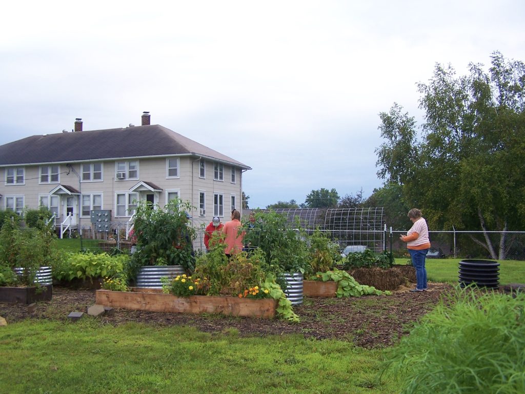 Garden plot with two-story Park View apartments building in the background. Three individuals standing and working in the garden beds.