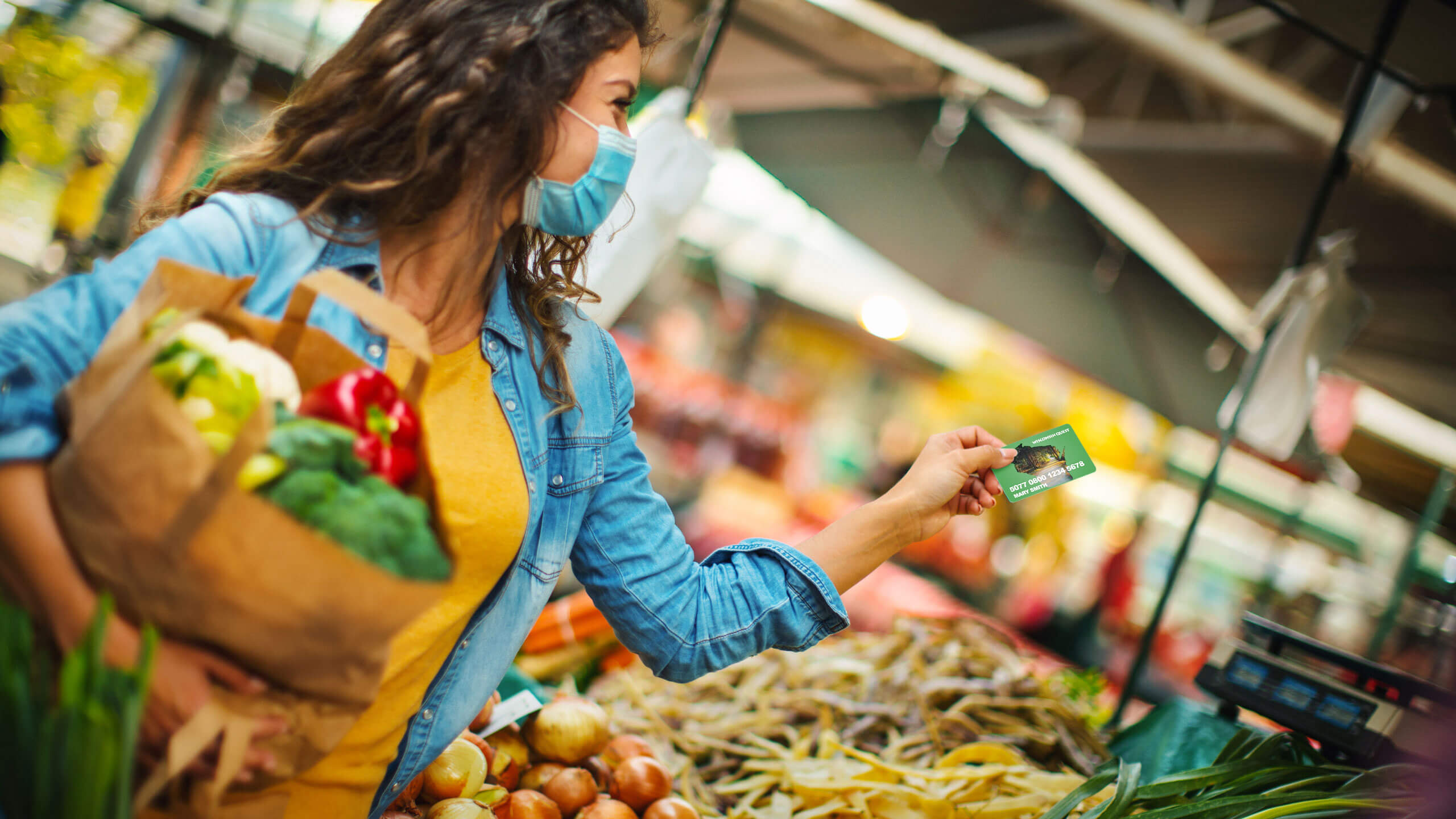 a woman holding a grocery bag filled with vegetables while handing her Quest card to the cashier 