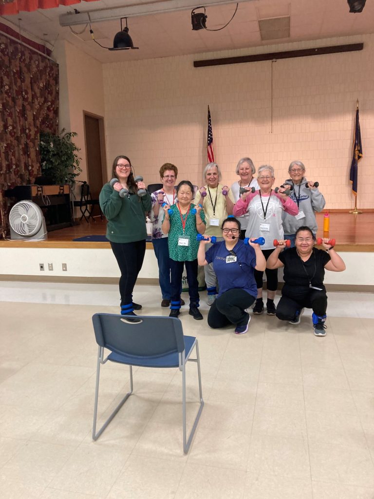 Educator Krystina, a young Hmong-American woman with glasses wearing a blue shirt and black pants, and holding blue hand-weights, kneels next to a Hmong StrongBodies participant holding red hand-weights. A row of 6 seniors stand behind Krystina and next to intern Morgan, a young European American woman in a green shirt and black pants holding grey hand-weights. 